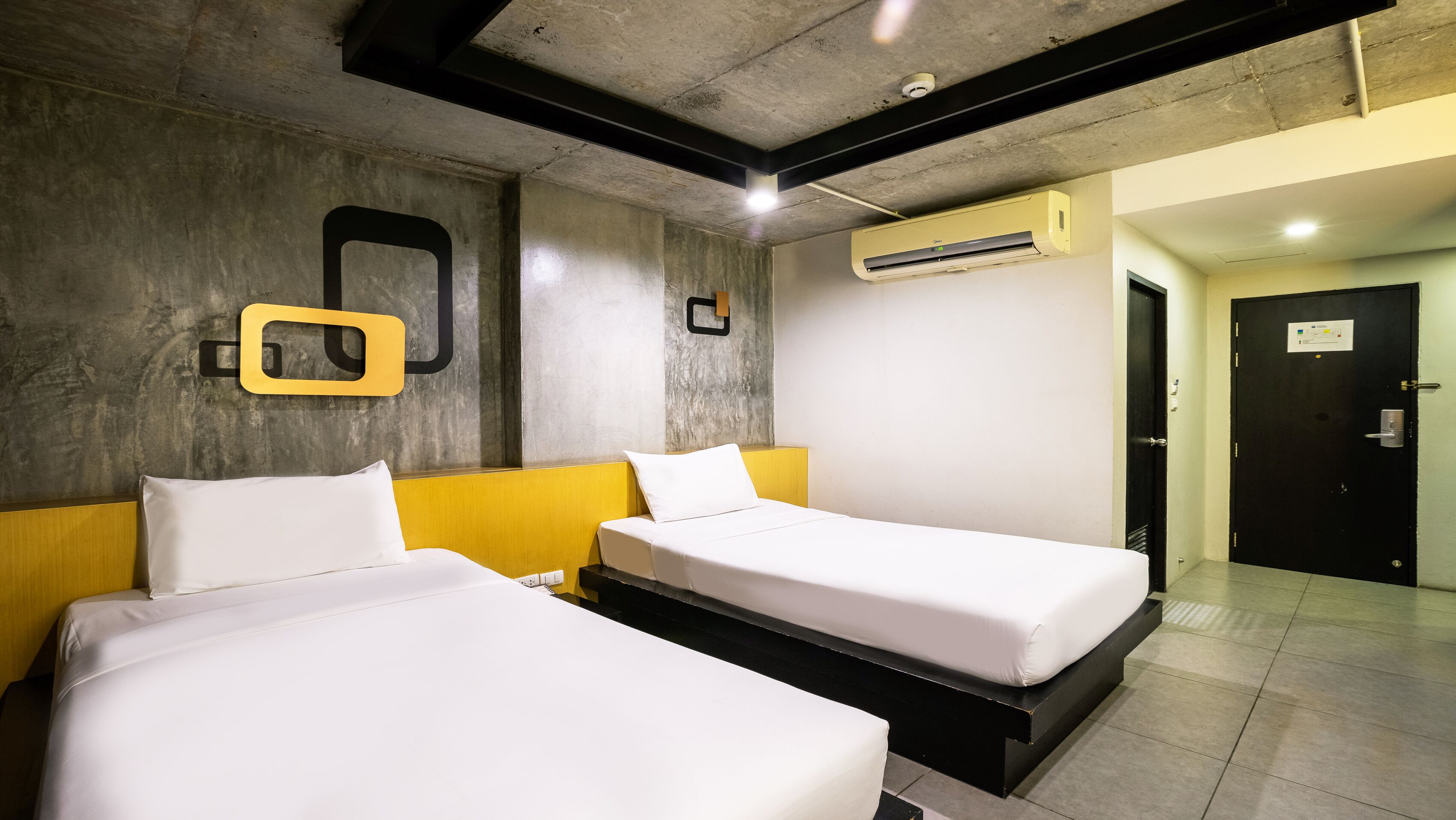 Habitación de hotel con dos camas, una pared de acento de concreto y otra de acento amarilla, además de un equipo de aire acondicionado.