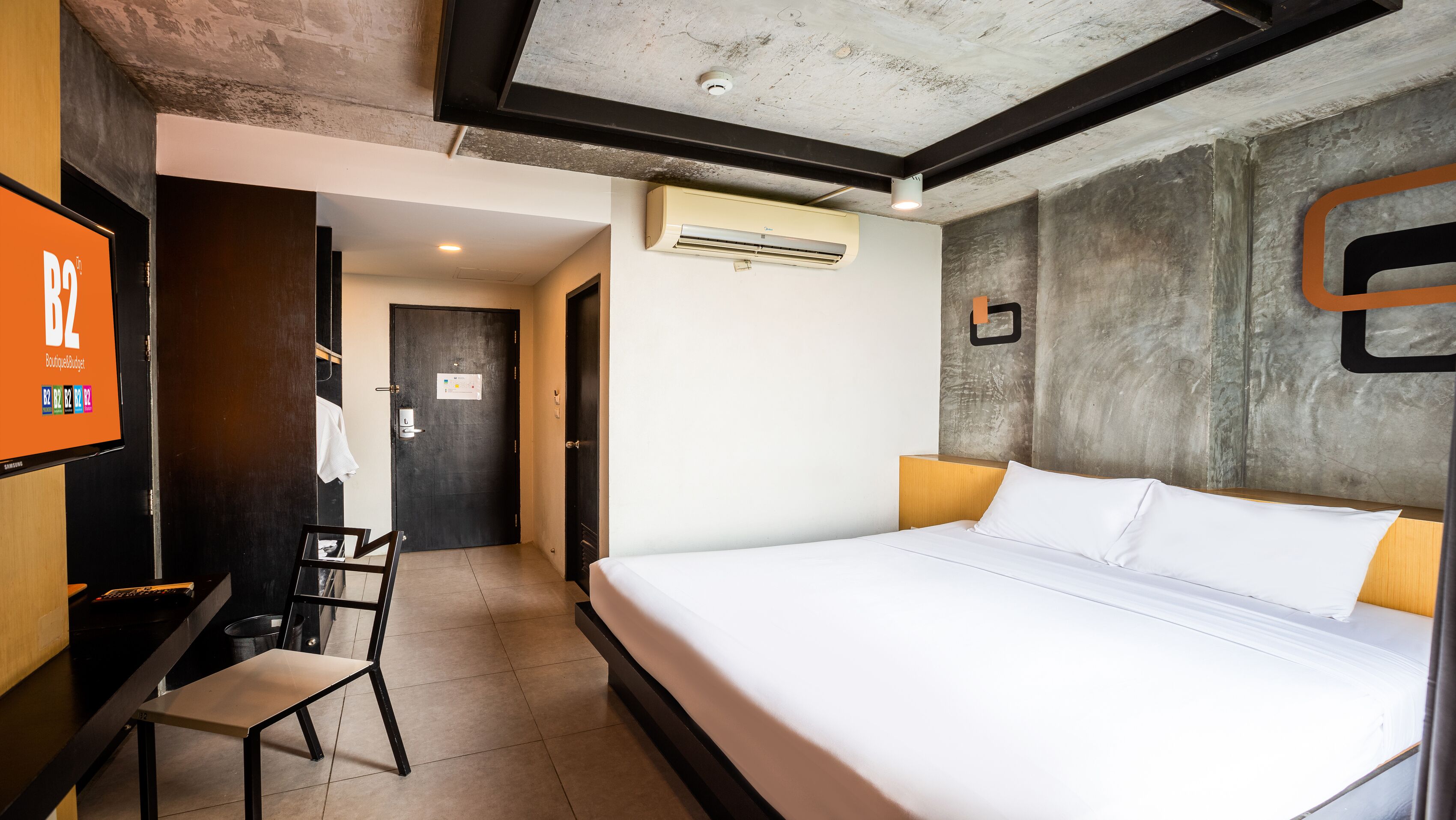 A hotel room with a bed, a desk with a chair, an air conditioning unit, and a TV displaying 'BoutiqueBudget'.