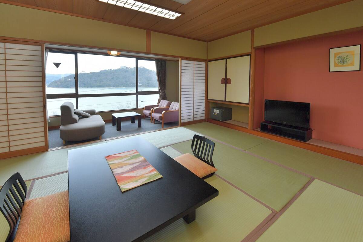 Ein traditioneller japanischer Raum mit Tatami-Böden, einem Tisch aus Tatami-Matten, einem Fernseher und Blick auf einen See.