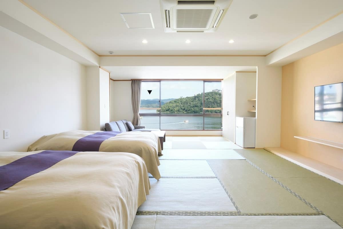 A hotel room with two beds, a large window with a view of a lake and hills, a small refrigerator, and a television on the wall.
