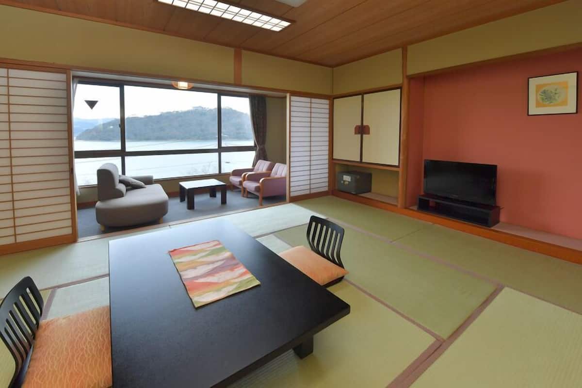 A room with tatami flooring, a tatami mat table, a sofa, a television, and a view of a lake.