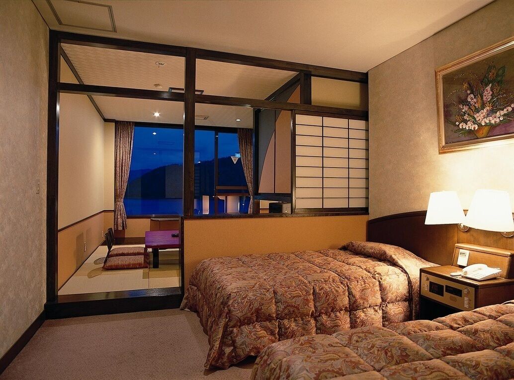 Ein Hotelzimmer mit einem Bett, einem Nachttisch, einer Lampe, einem Fenster mit Blick und einem Bild an der Wand.
