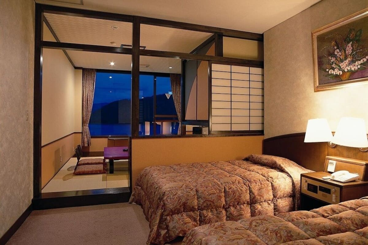 A hotel room with a bed, a nightstand, a lamp, a window with a view, and a painting on the wall.