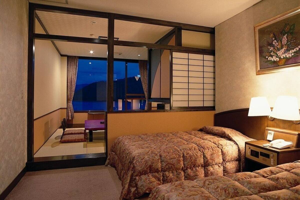 Ein Hotelzimmer mit einem Bett, einem Nachttisch, einer Lampe, einem Fenster mit Blick und einem Bild an der Wand.