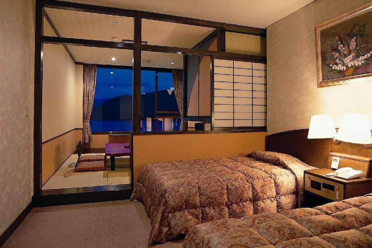 A hotel room with a large window, a bed with patterned bedding, a nightstand with a lamp, and a painting on the wall.