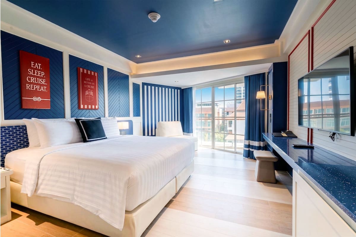 A modern hotel room with a large bed, a desk, a view of the city, and two red signs on the wall.