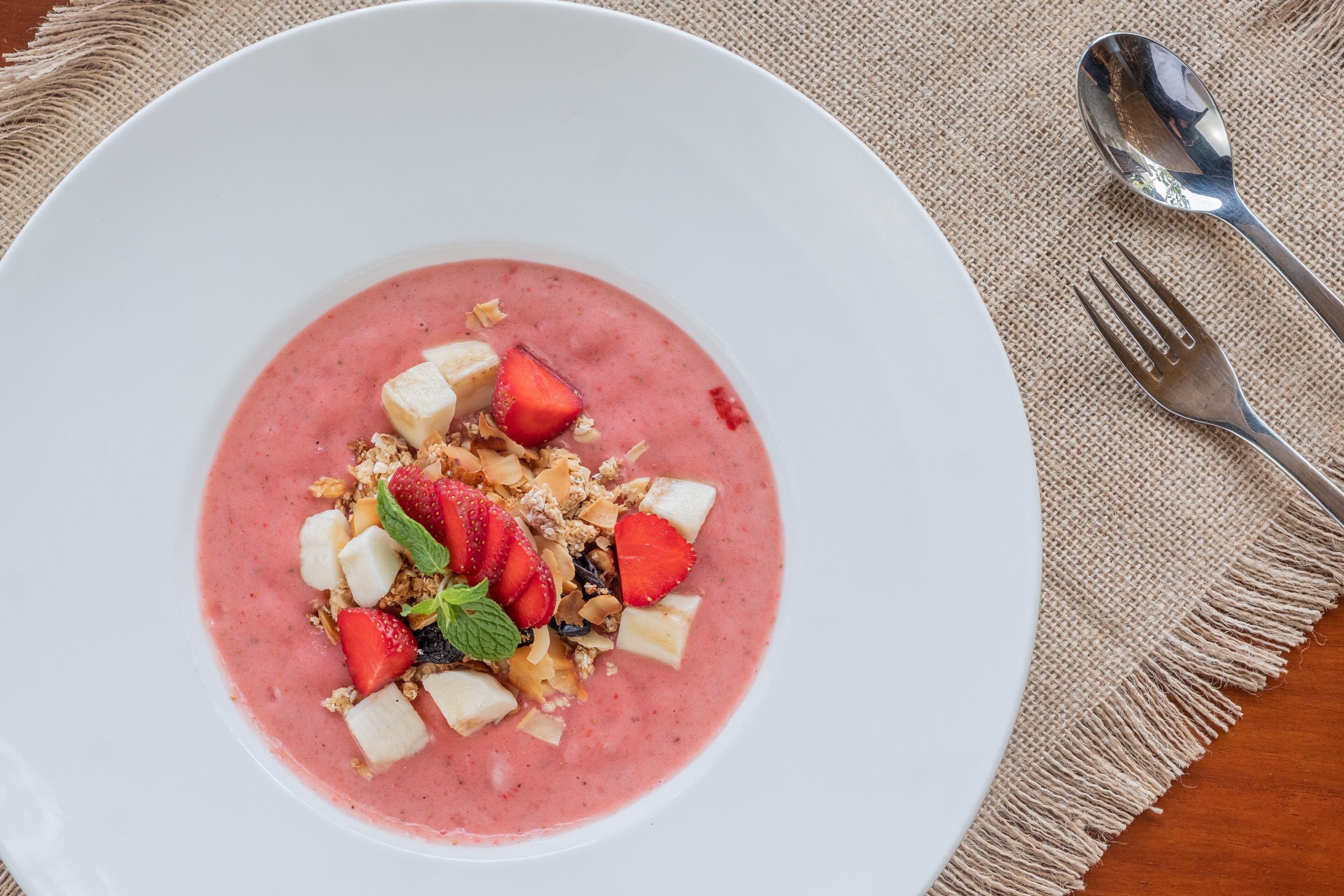 En hvid tallerken med en jordbær smoothie pyntet med jordbær, banan, granola og et mynteblad.