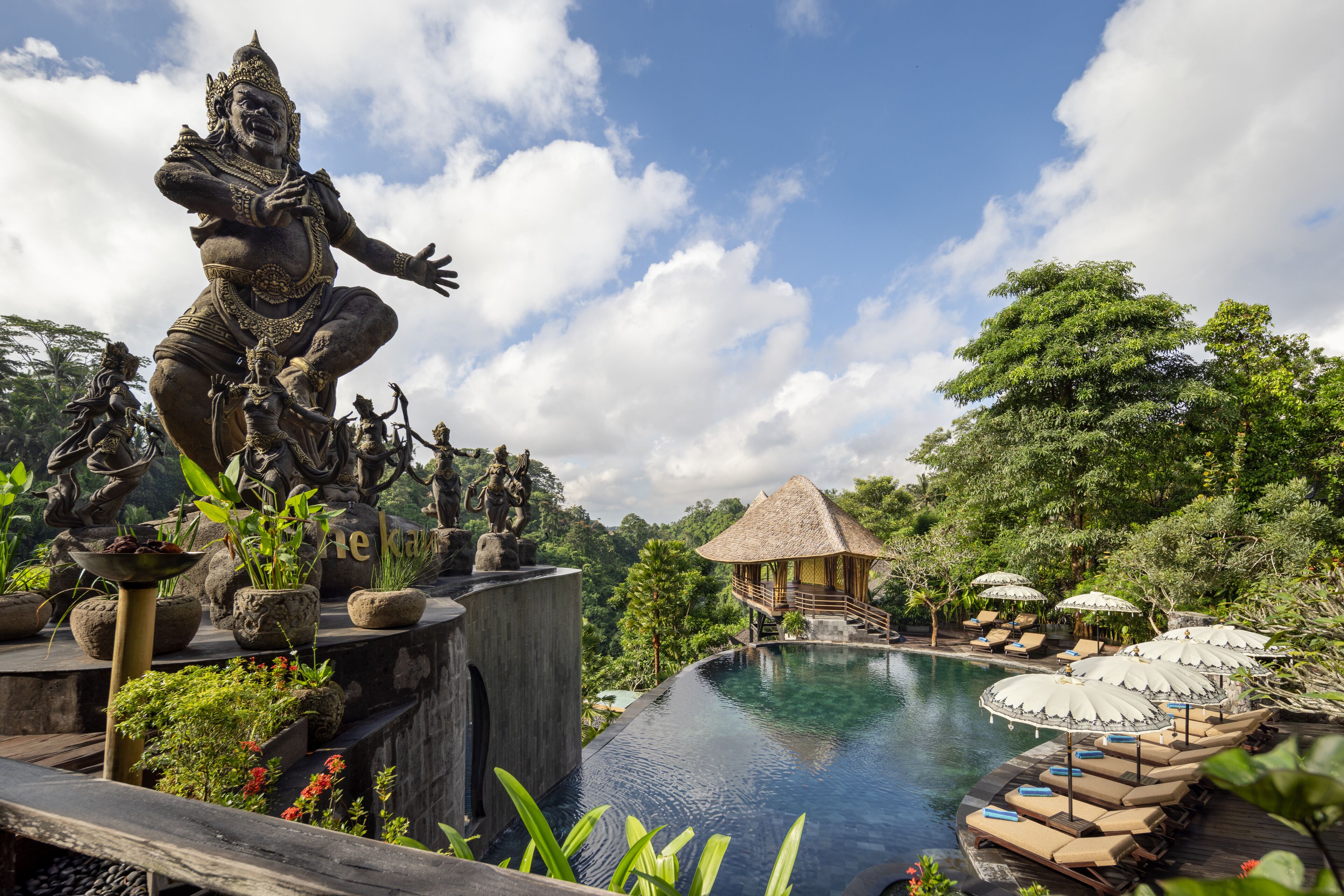 A pool area with statues, lounge chairs, and a thatched-roof structure.
