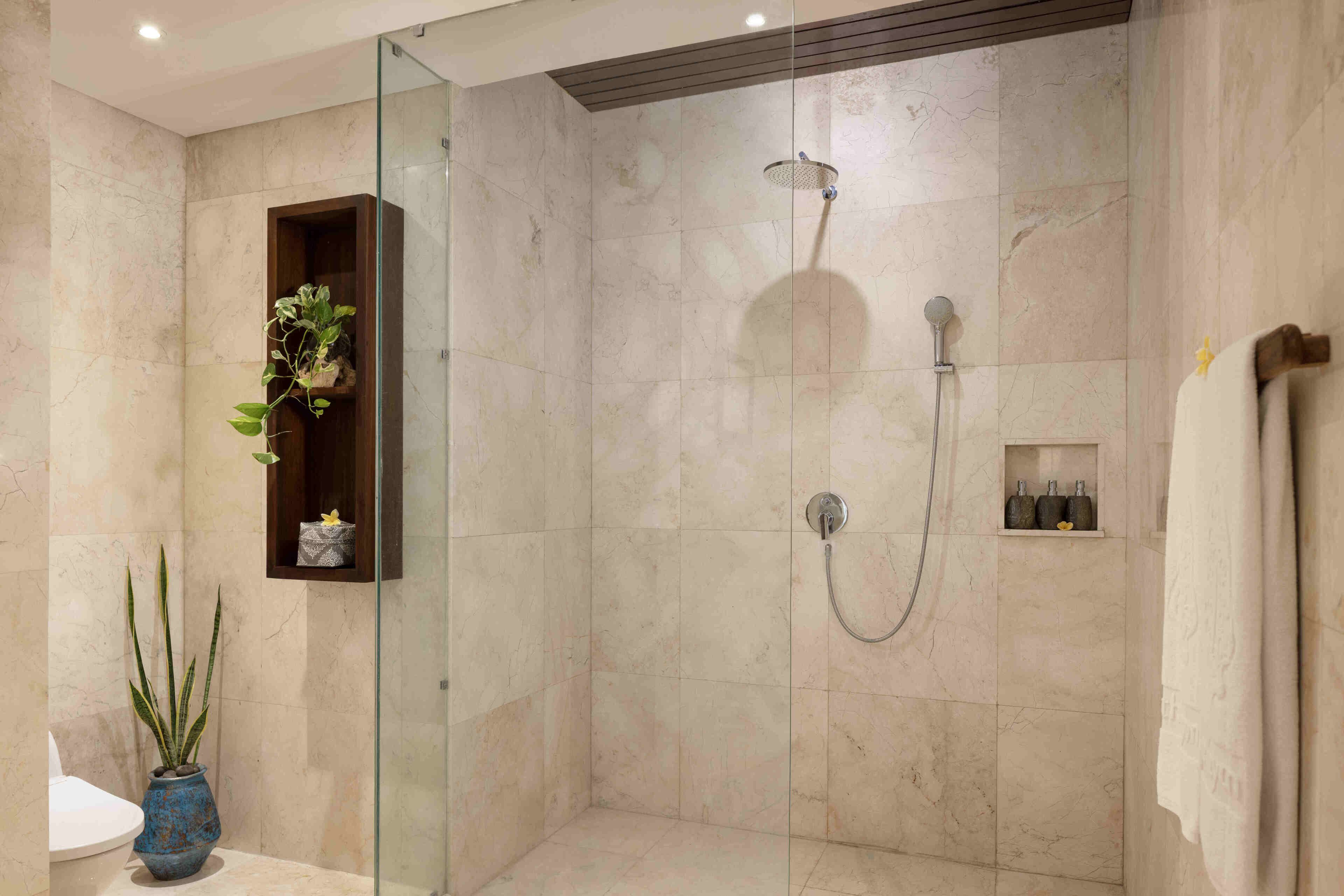 Un baño moderno con ducha de vidrio, azulejos de mármol, una planta en maceta y un pequeño estante con artículos de aseo.