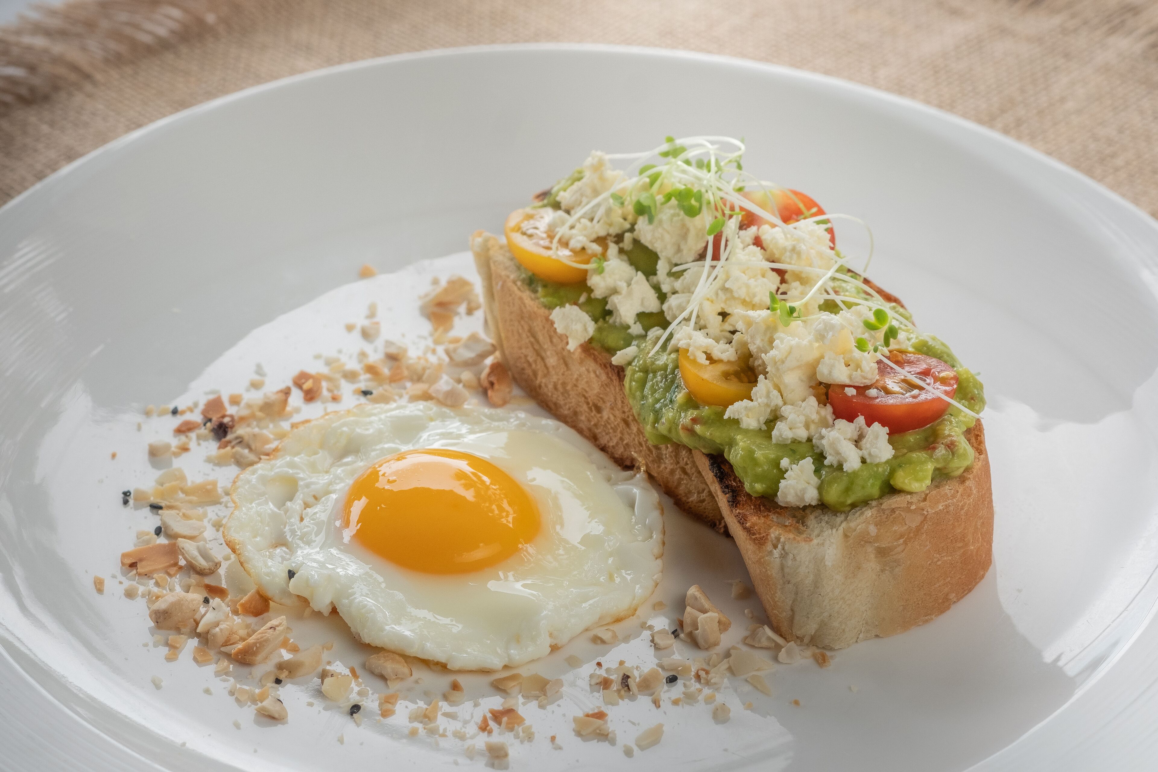 Een bord met een gebakken ei, avocado-toast en een sneetje brood belegd met tomaten, kaas en kiemgroenten.