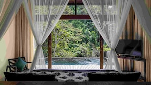 Un dormitorio con un ventanal grande, una cama, un televisor y vistas al verdor exterior.