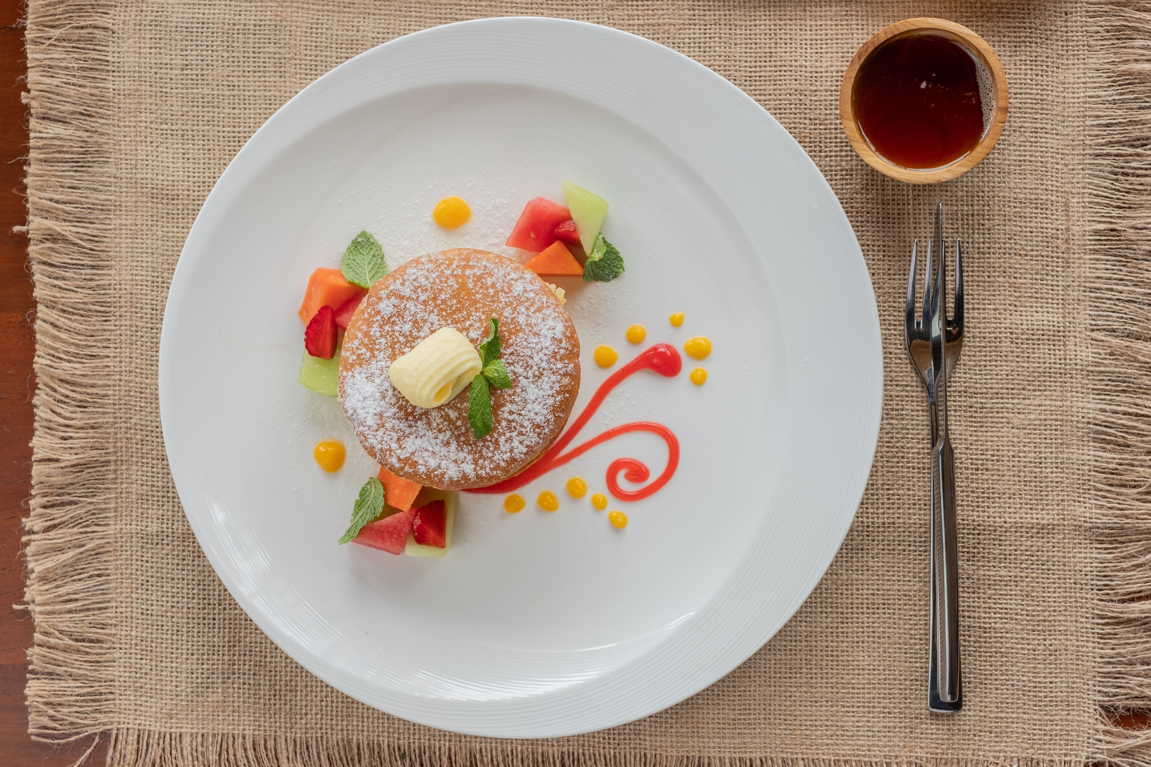 Postre en un plato blanco, con un tenedor y un recipiente pequeño de almíbar sobre un mantel de arpillera.