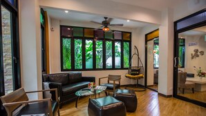 A modern living room with a leather sofa, wooden chairs, a glass coffee table, and a ceiling fan. Large windows with green foliage outside are visible. - Tee Pak Dee Resident Phuket (Phuket)