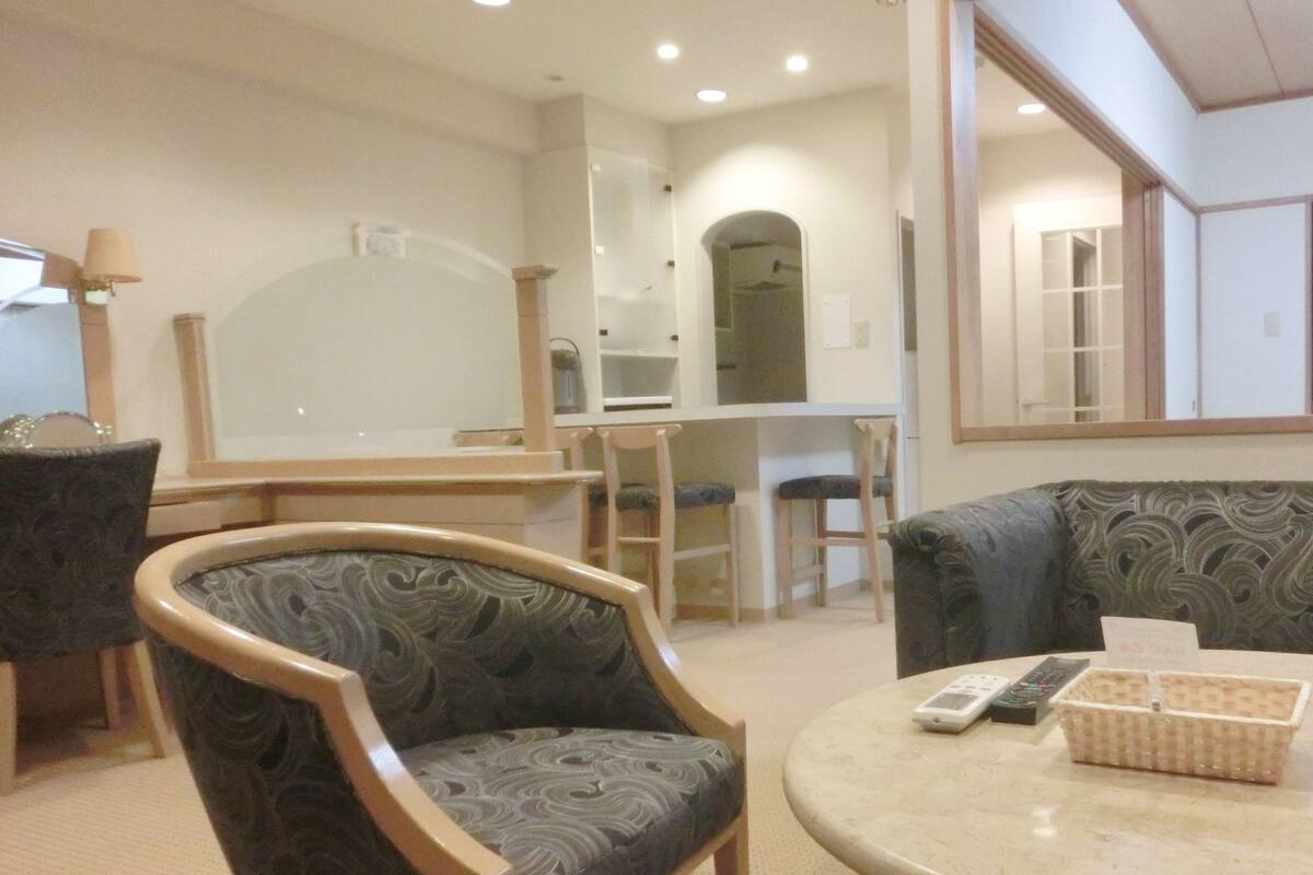 Sala de estar com sofá, cadeiras, uma mesa e área de cozinha ao fundo.