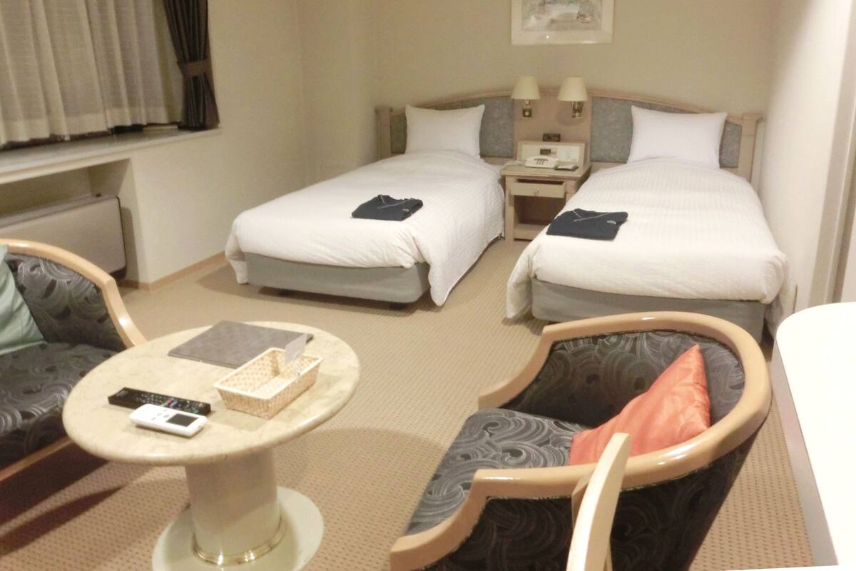 Quarto de hotel com duas camas, uma mesinha, duas cadeiras e uma mesa de cabeceira com luminárias.