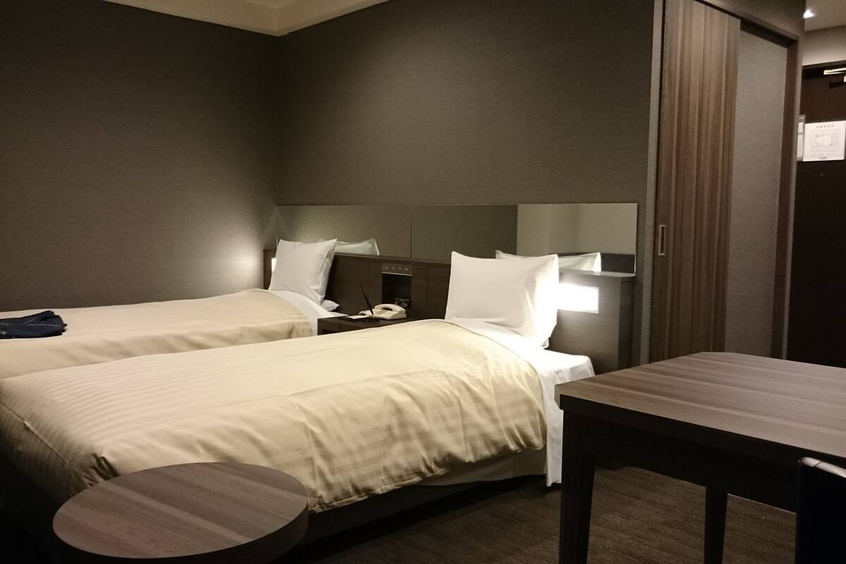 Quarto de hotel com duas camas, uma escrivaninha, uma cadeira, um telefone e um espelho.
