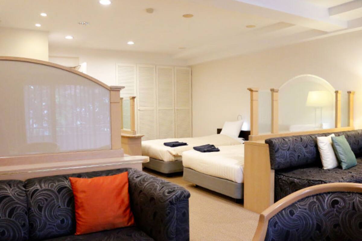 Quarto de hotel com duas camas, um sofá, um espelho e uma janela grande.