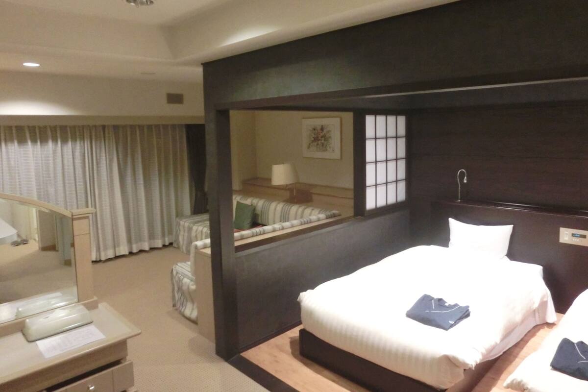 Quarto de hotel com uma cama grande, uma mesa de cabeceira, um espelho, uma janela com persianas e vista para o corredor.