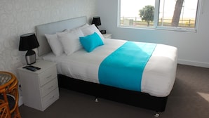 A neatly made bed with white and blue bedding, a bedside table with a lamp, and a view of the outdoors through a window. - Beach Hotel (Burnie)