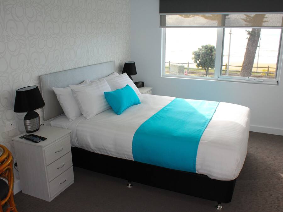 A neatly made bed with white and blue bedding, a bedside table with a lamp, and a view of the outdoors through a window.