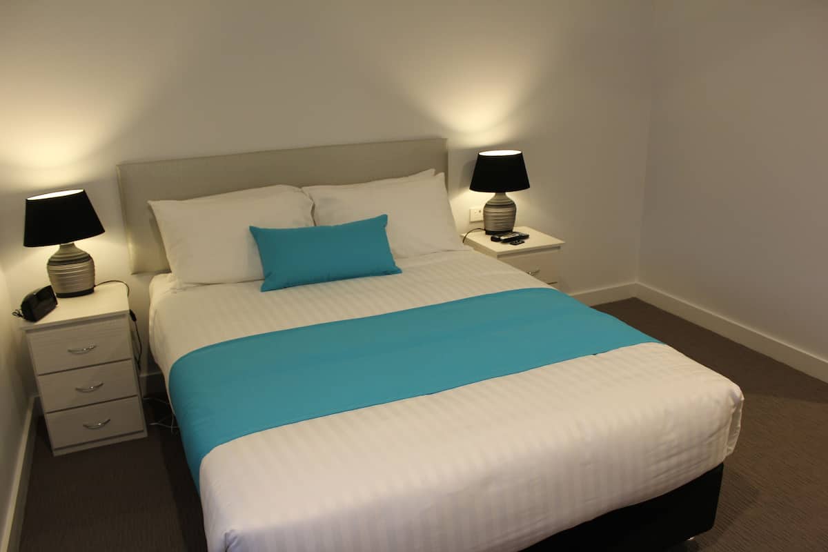 Ein ordentlich bezogenes Bett mit blauem Zierkissen, zwei Nachttische mit Lampen und eine Kommode.