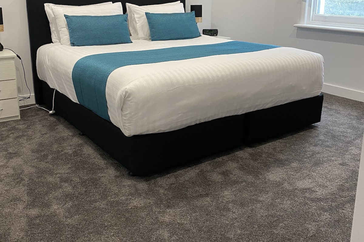 Ein ordentlich bezogenes Bett mit schwarzem Kopfteil, weiß-blauer Bettwäsche und einem grauen Teppichboden.