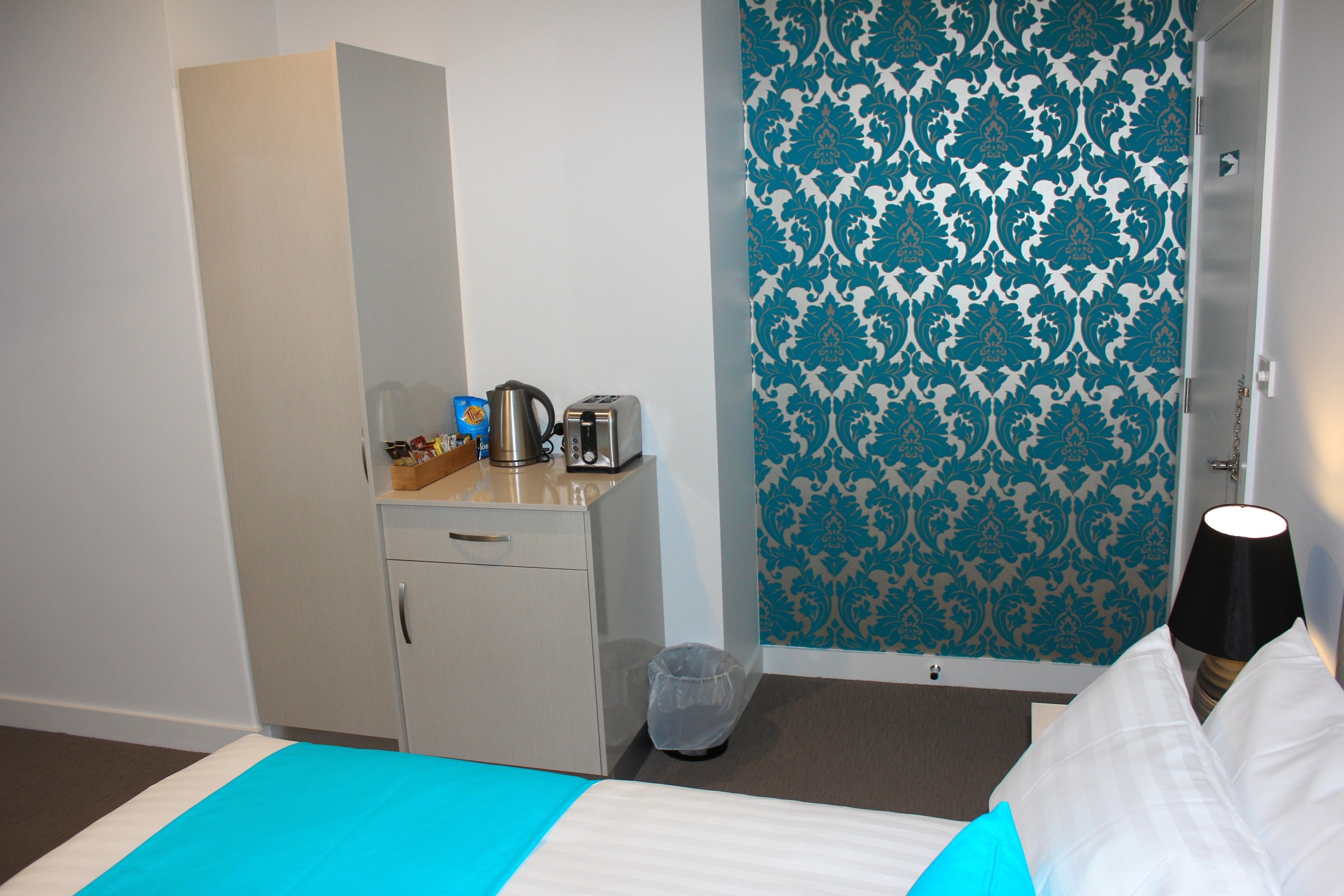 A hotel room with a bed, a small desk with a kettle and toaster, a trash bin, and a patterned wall.