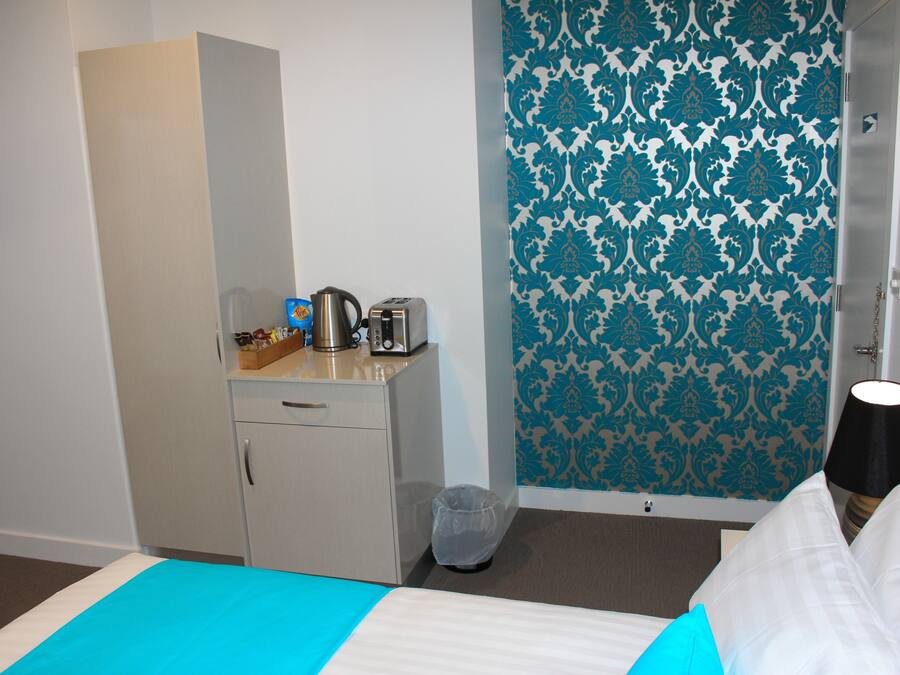 Una habitación de hotel con una cama, un pequeño escritorio con hervidor y tostadora, un cesto de basura y un papel tapiz estampado.