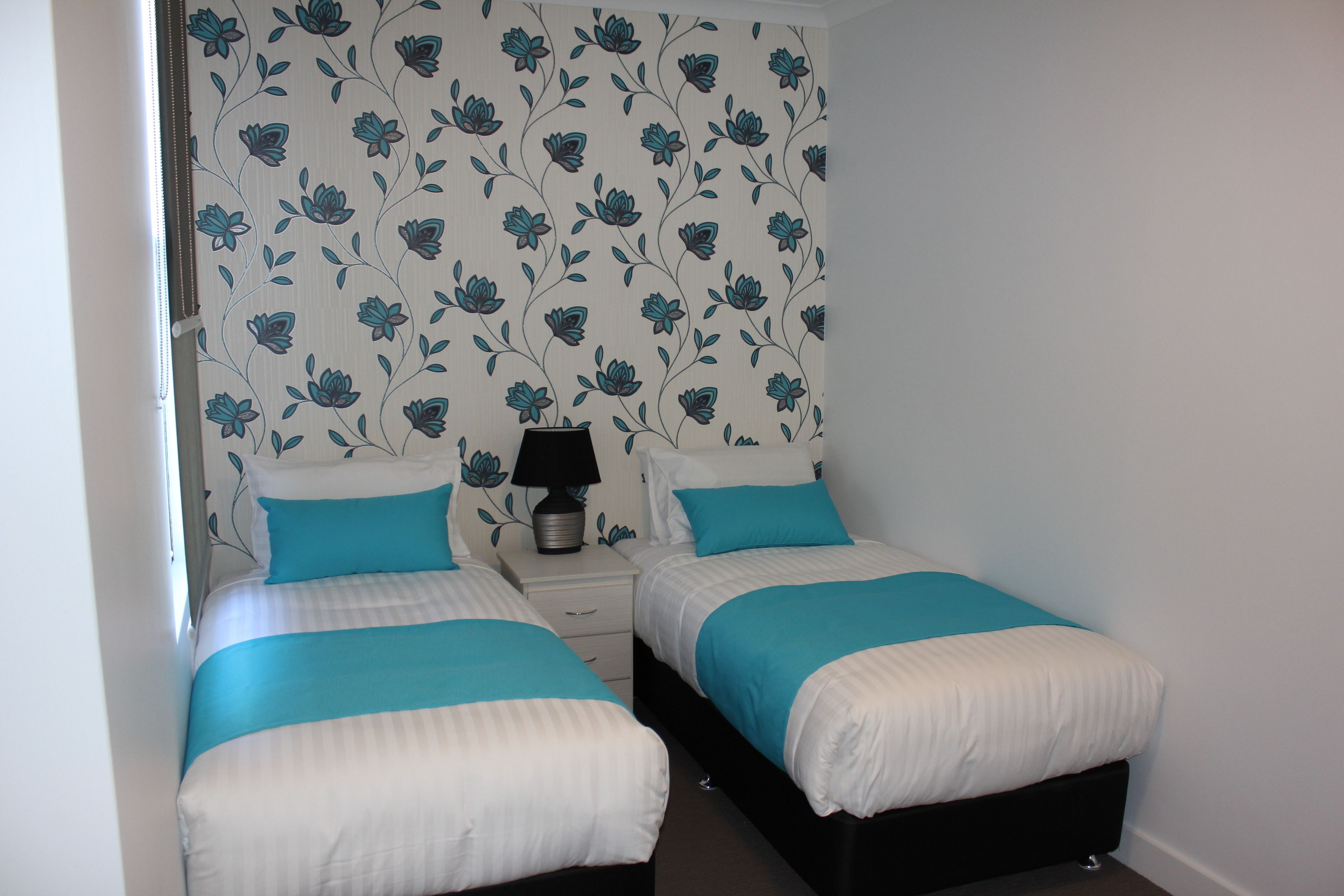 A room with two beds, each with a blue and white bedspread, a bedside table with a lamp, and floral wallpaper.
