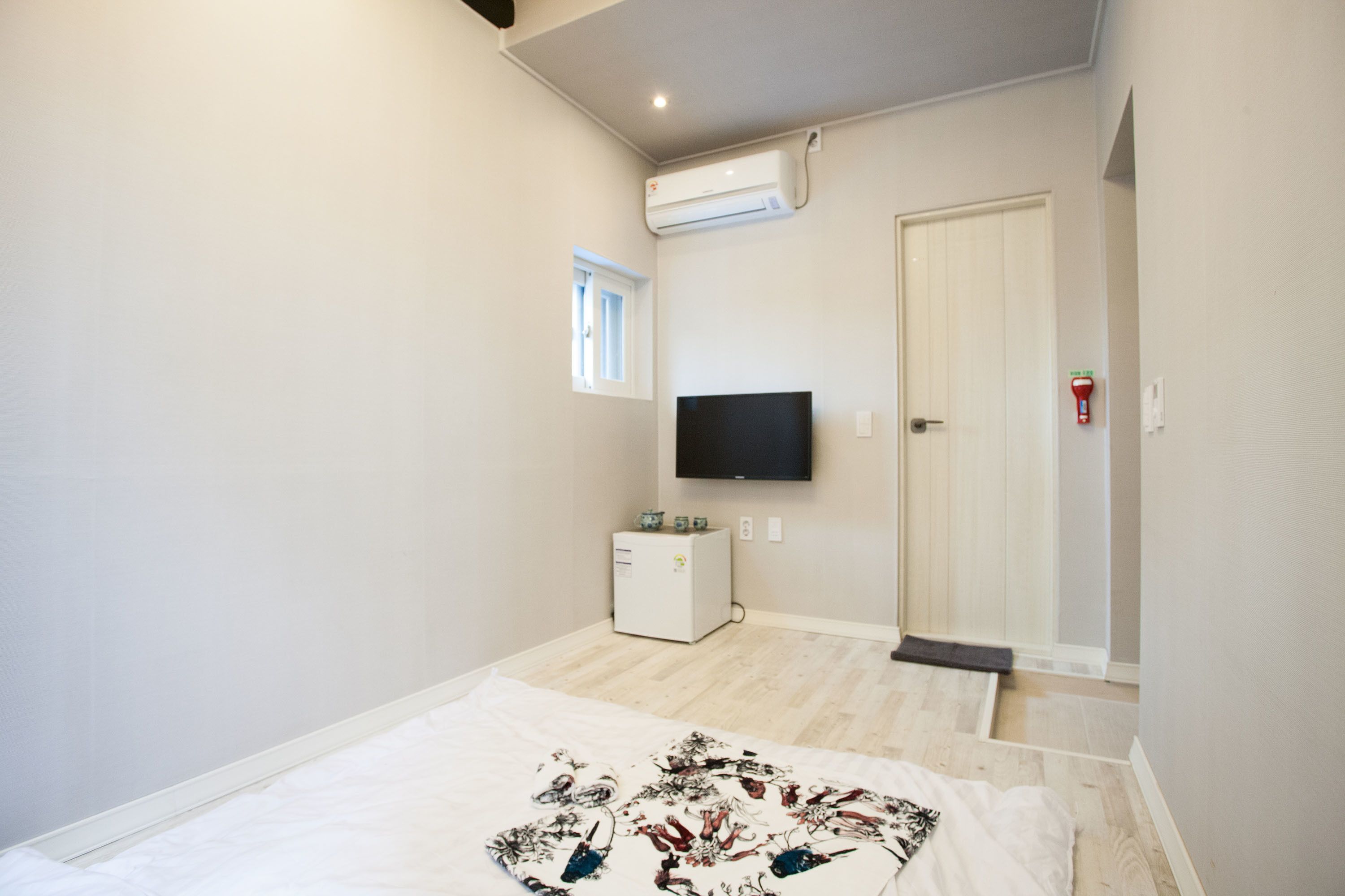 Un dormitorio con cama, televisión montada en la pared, refrigerador pequeño y una puerta que da a otra habitación.