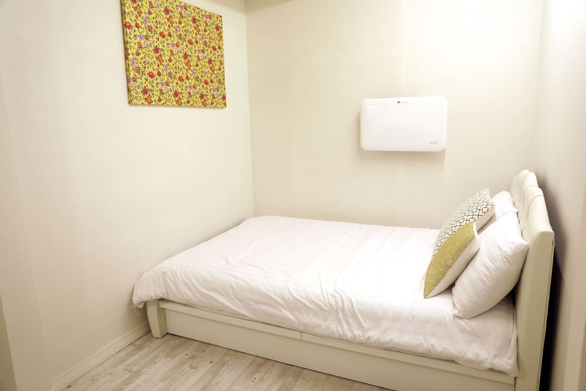 Un petit lit bien fait, avec une literie blanche et un coussin décoratif, un climatiseur fixé au mur et une œuvre d’art aux motifs floraux accrochée au mur.