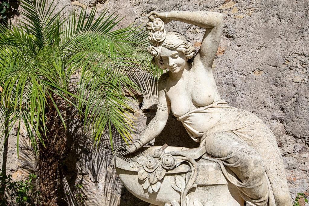 Una scultura in pietra raffigura una donna sdraiata con un fiore tra i capelli, accanto a una pianta di palma.