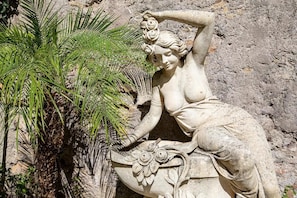 Une sculpture en pierre représentant une femme allongée, avec une fleur dans les cheveux, placée à côté d’un palmier.