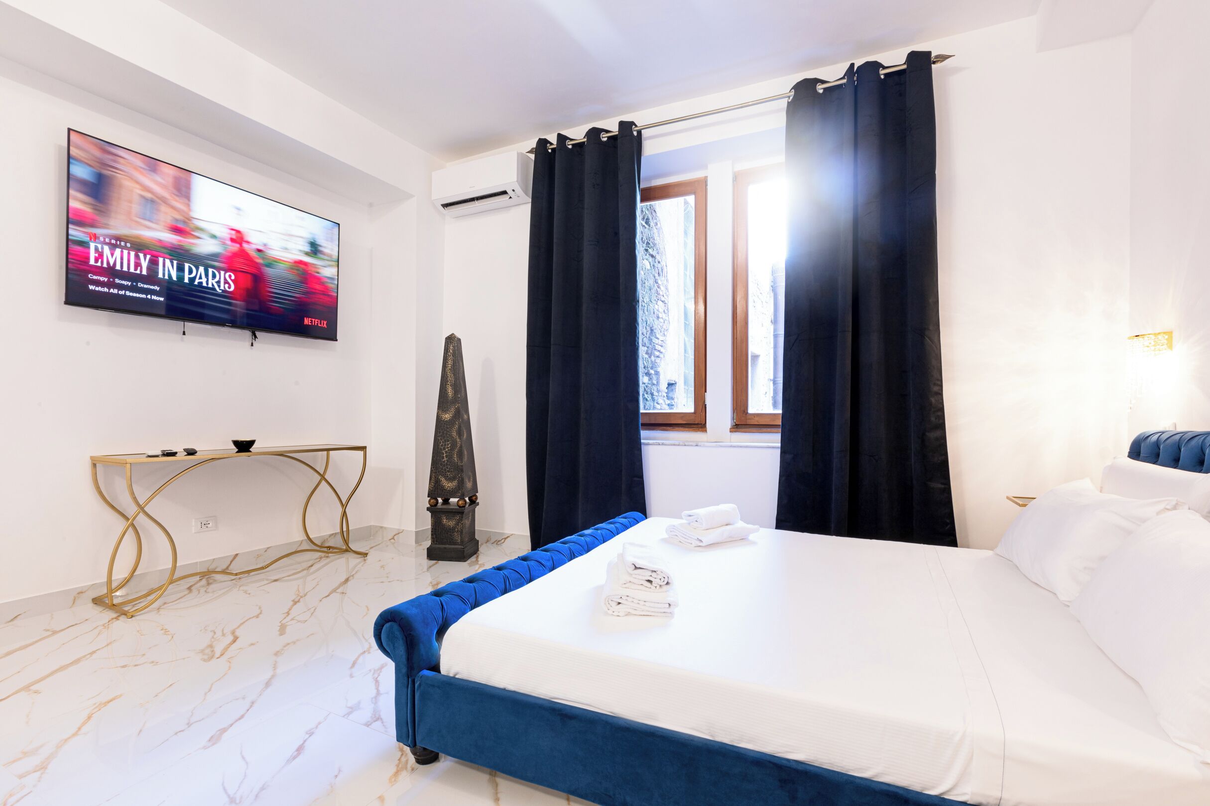 A hotel room with a bed, a TV displaying 'Emily in Paris', a console table, and a window with curtains.