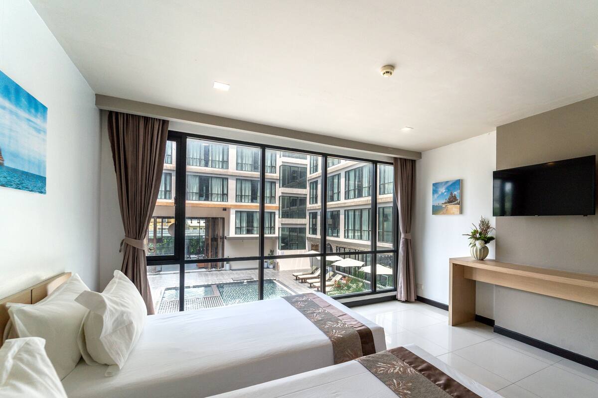 A hotel room with a large window, a bed, a bedside table, a TV, and a view of the building's exterior.