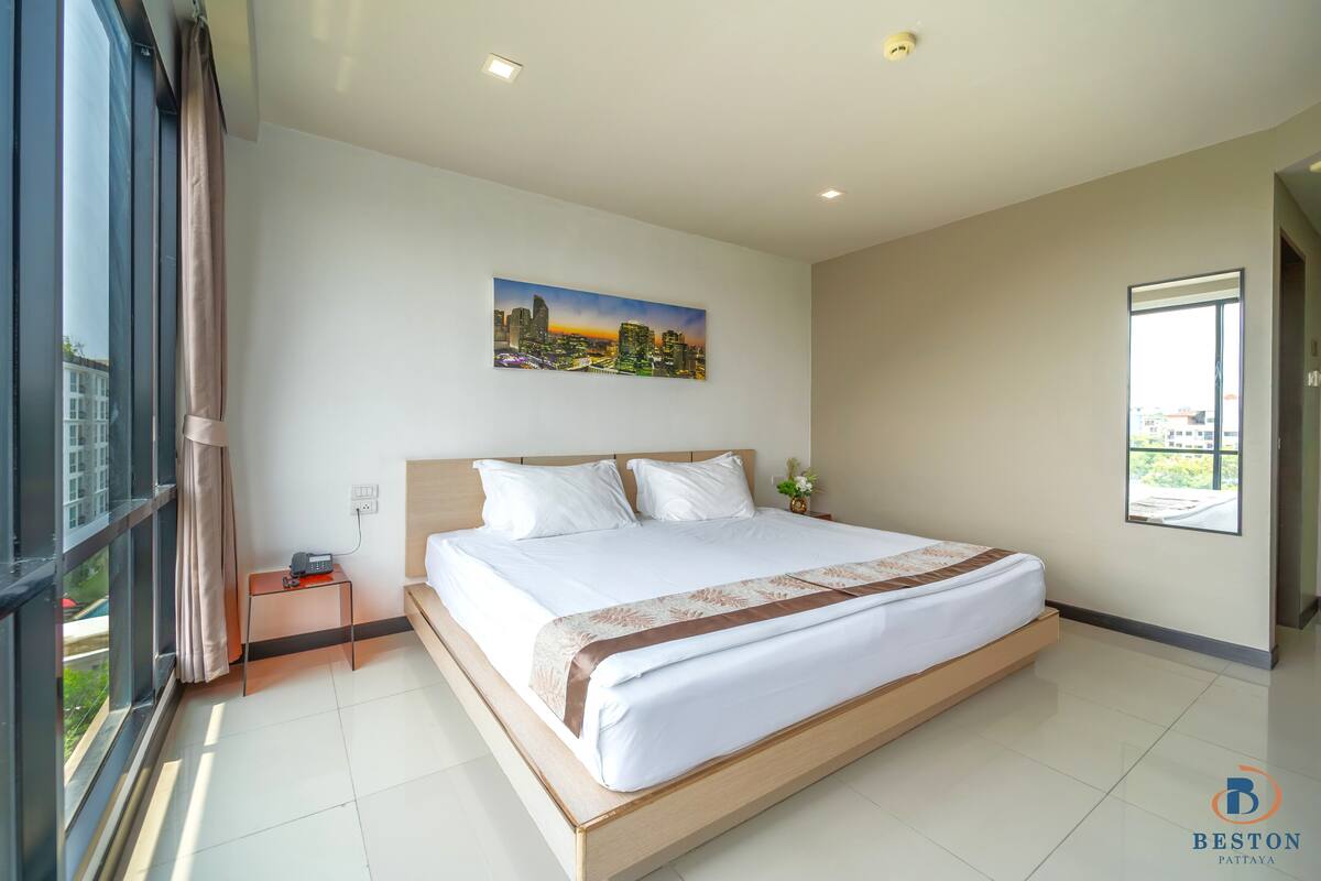 A modern hotel room with a large bed, a painting of a cityscape, a nightstand, and a window with a view.