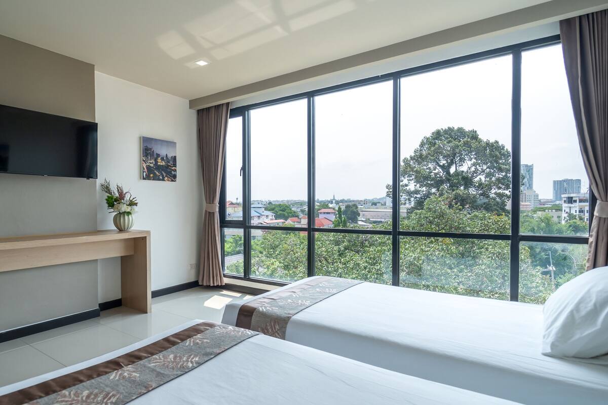 A hotel room with a large window, a flat-screen TV mounted on the wall, a desk with a chair, and two beds.