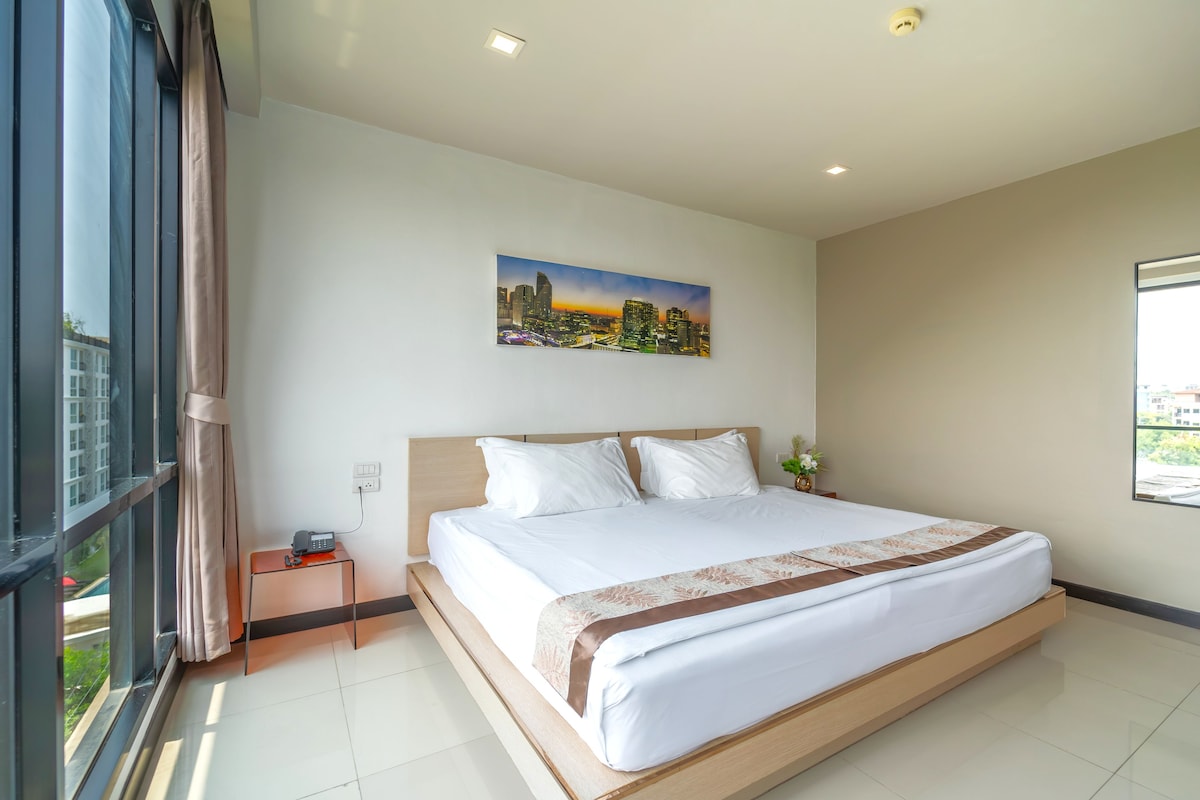 A modern hotel room with a large bed, a painting of a cityscape, a nightstand, and a window with a view.