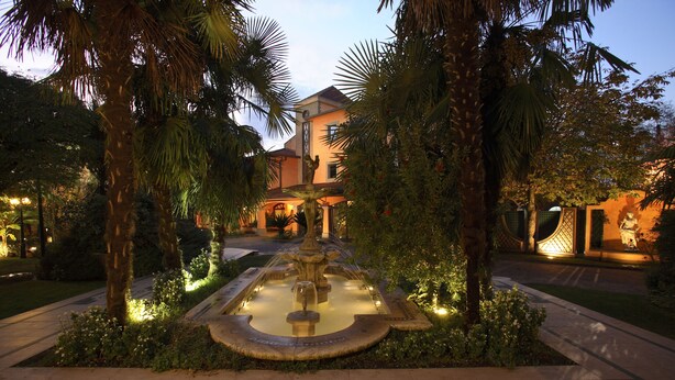 A well-lit courtyard with a central fountain, surrounded by palm trees and a building with a traditional architectural style. - Hotel Borgo Antico (Ceprano)