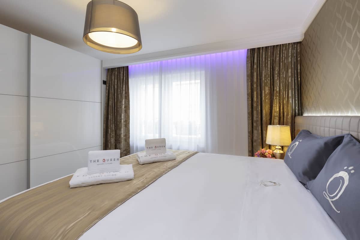 A hotel room with a large bed, a wooden headboard, a nightstand with a lamp, and a window with curtains.