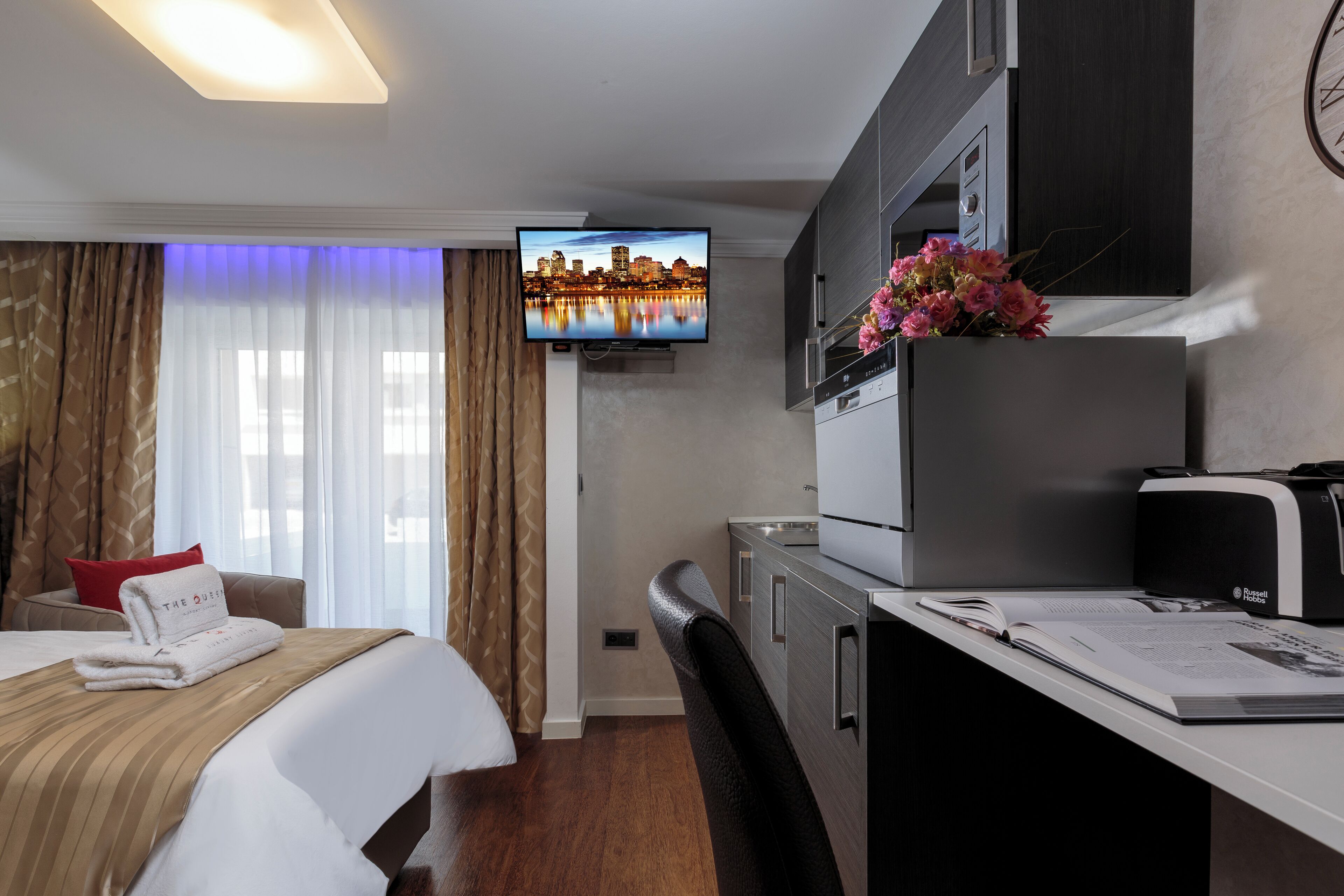 Habitación de hotel con una cama, un escritorio con computadora, un televisor mostrando un paisaje urbano y un reloj en la pared.