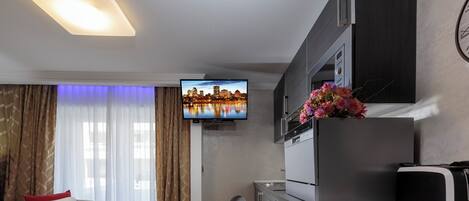 Habitación de hotel con una cama, un escritorio con computadora, un televisor mostrando un paisaje urbano y un reloj en la pared.