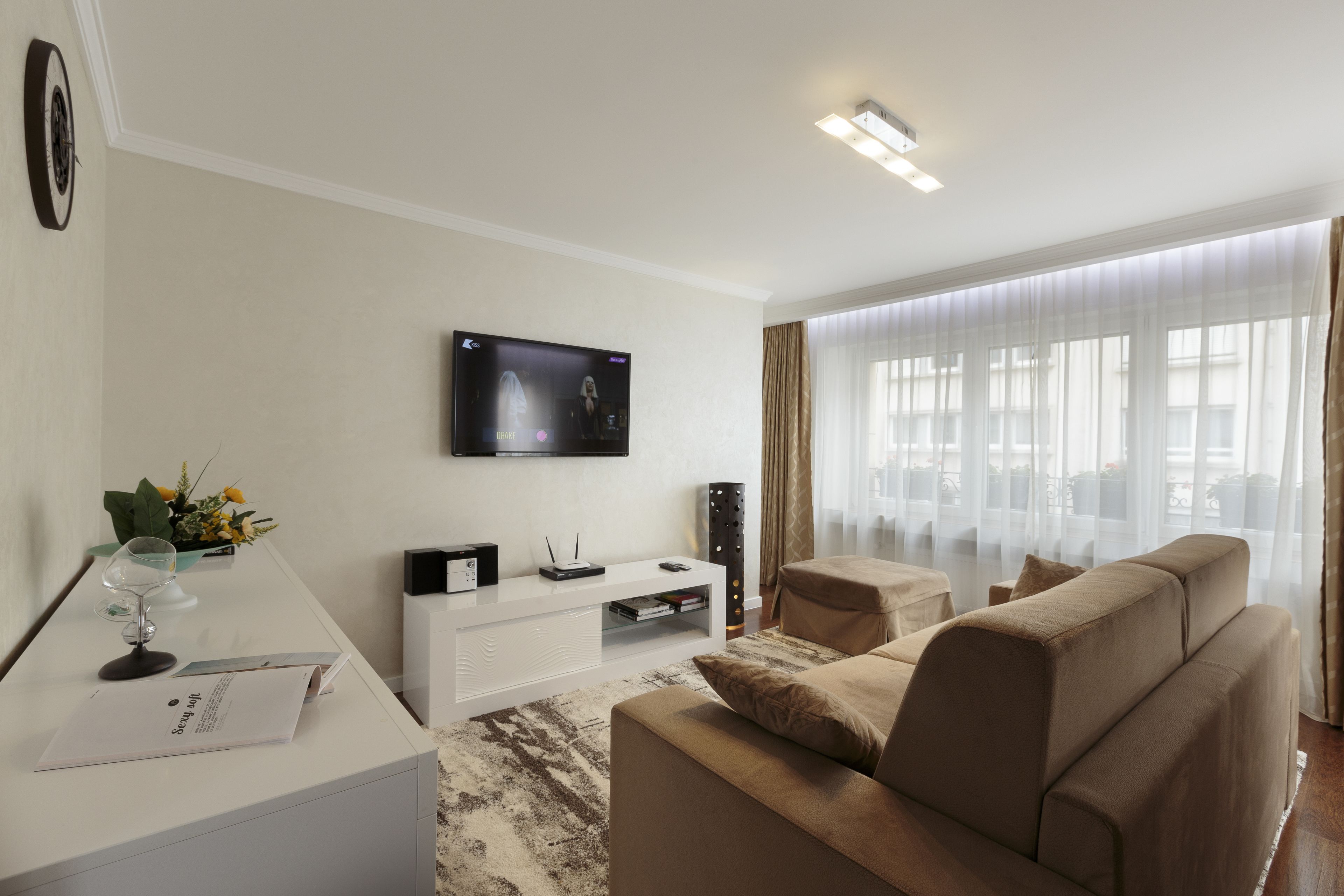 Un salón moderno con un sofá, un televisor de pantalla plana montado en la pared, un mueble blanco para entretenimiento y un ventanal con cortinas de gasa.