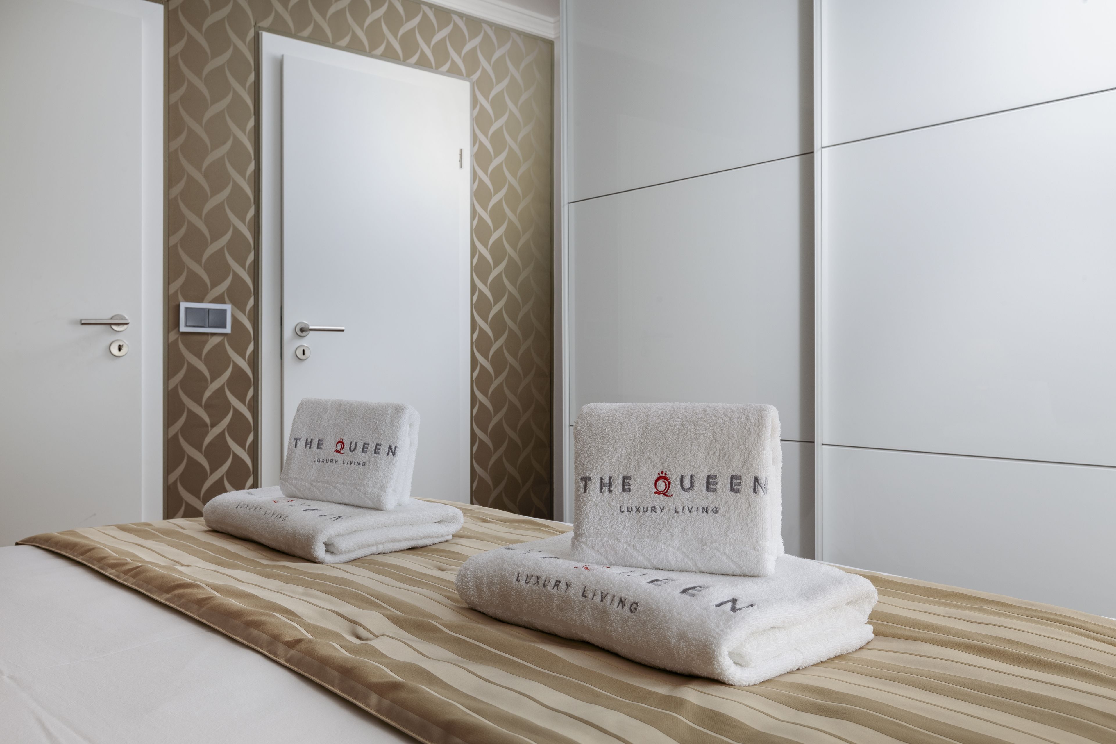 Une chambre d’hôtel avec un lit, deux serviettes portant la marque « The Queen Luxury Living », une porte et un mur décoré d’un motif.