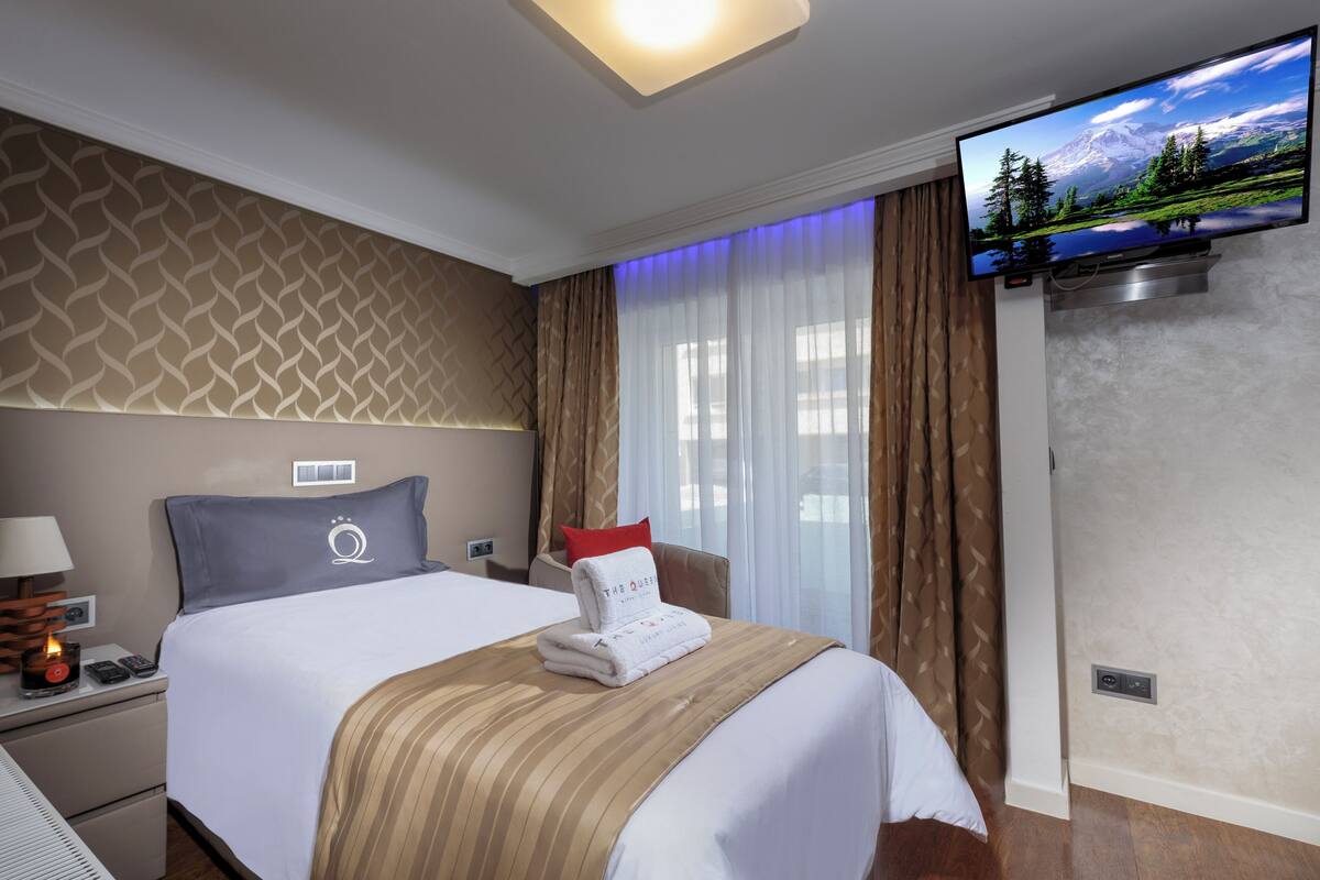 A hotel room with a bed, a flat-screen TV mounted on the wall, a nightstand with a lamp, and a window with curtains.