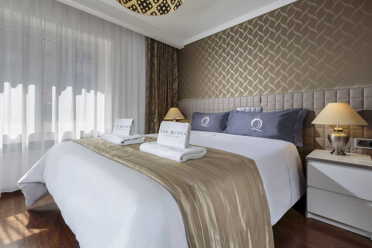 A hotel room with a large bed, two bedside tables, a nightstand, a patterned wall, and sheer curtains.