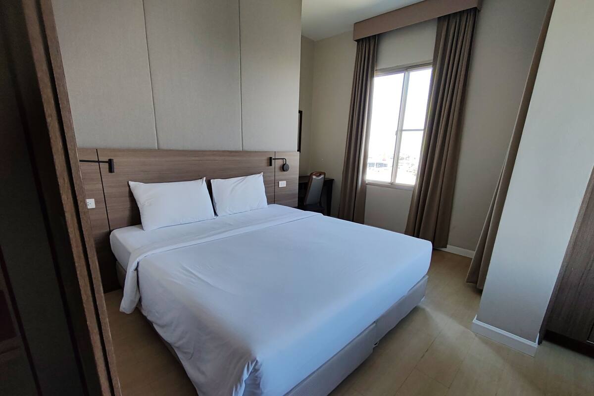 Uma cama bem arrumada em quarto de hotel, com cabeceira de madeira, mesa de cabeceira e janela com cortinas.