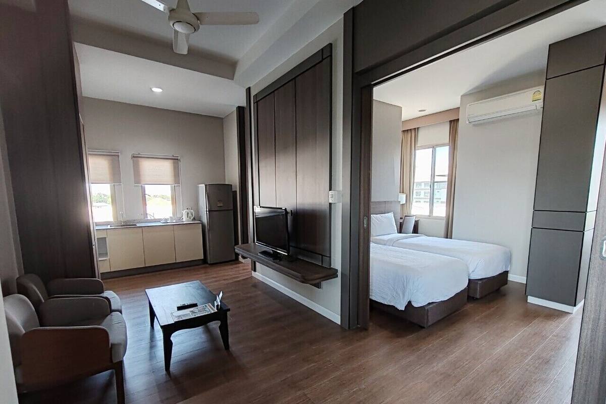 Quarto de hotel moderno com uma cama grande, uma televisão na parede, uma pequena cozinha e uma área de estar com uma mesa de centro.