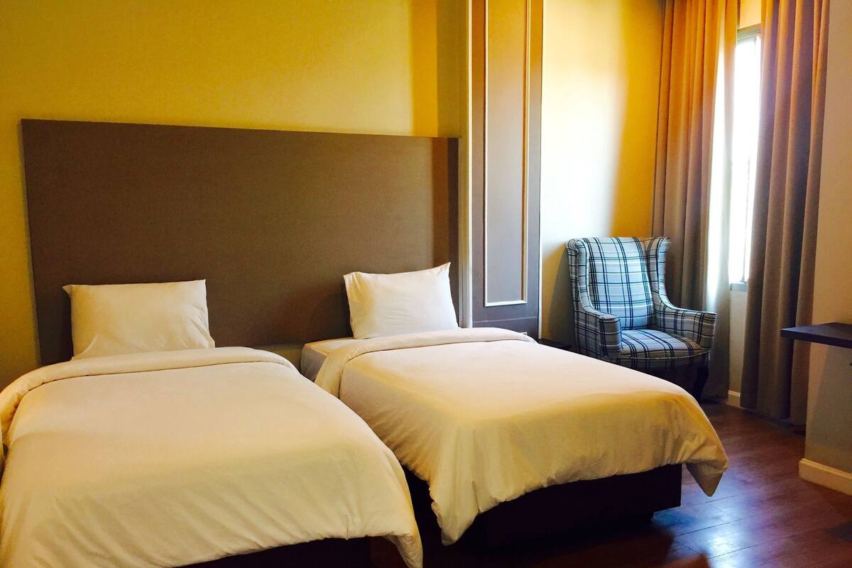 Quarto de hotel com duas camas, uma cadeira e uma janela com cortinas.