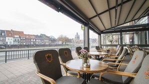 A covered outdoor seating area with wicker chairs and tables, overlooking a cityscape with buildings and a river. - Hotel en Grand Café de Pauw (Roermond)